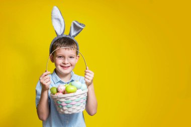 Paskalya gününde tavşan kulağı takan ve elinde boyalı yumurtalarla sepet tutan şirin bir çocuk. Mutlu çocuk elinde Paskalya sepeti tutuyor..