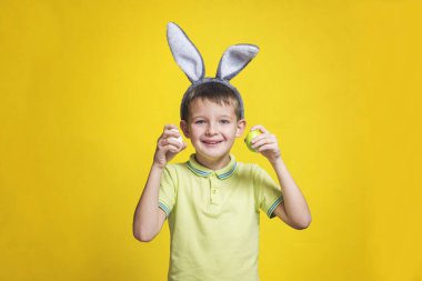 Paskalya tavşanı kulaklı şirin bir çocuk iki renkli paskalya yumurtası tutuyor. Tavşan kulaklı ve sarı arka planda paskalya yumurtaları olan bir çocuk..