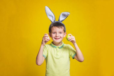 Paskalya tavşanı kulaklı şirin bir çocuk iki renkli paskalya yumurtası tutuyor. Tavşan kulaklı ve sarı arka planda paskalya yumurtaları olan bir çocuk..