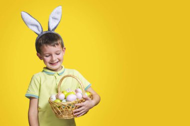 Paskalya gününde tavşan kulağı takan ve elinde boyalı yumurtalarla sepet tutan şirin bir çocuk. Mutlu çocuk elinde Paskalya sepeti tutuyor..