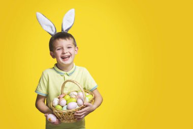 Paskalya gününde tavşan kulağı takan ve elinde boyalı yumurtalarla sepet tutan şirin bir çocuk. Mutlu çocuk elinde Paskalya sepeti tutuyor..