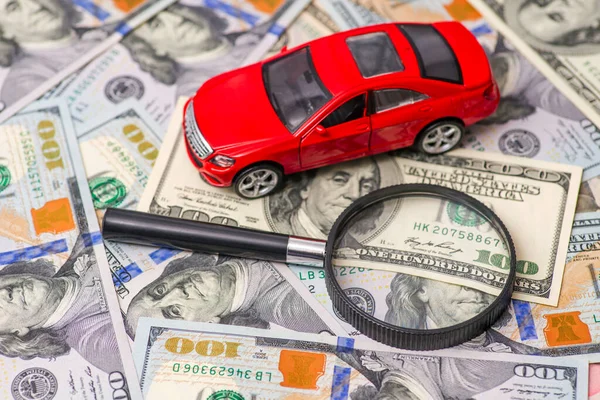 Magnifying glass and toy red car on dollar bills background. Saving money for car. Concept for advertising loan, collateral, pawnshop, car rental.