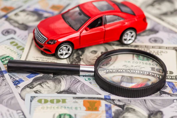 Magnifying glass and toy red car on dollar bills background. Saving money for car. Concept for advertising loan, collateral, pawnshop, car rental.