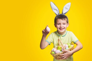 Paskalya tavşanı kulağı takan mutlu bir gülen çocuk sarı arka planda boyalı yumurtalarla dolu bir sepet tutuyor. Uzayı kopyala Paskalya Günü.