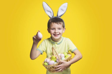 Paskalya tavşanı kulağı takan mutlu bir gülen çocuk sarı arka planda boyalı yumurtalarla dolu bir sepet tutuyor. Uzayı kopyala Paskalya Günü.