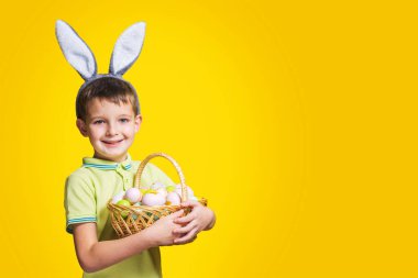 Paskalya tavşanı kulağı takan mutlu bir gülen çocuk sarı arka planda boyalı yumurtalarla dolu bir sepet tutuyor. Uzayı kopyala Paskalya Günü.