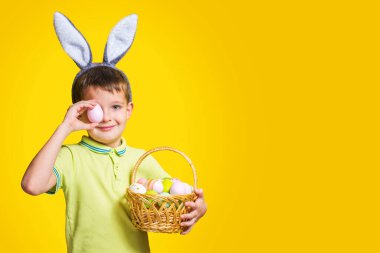 Paskalya tavşanı kulağı takan mutlu bir gülen çocuk sarı arka planda boyalı yumurtalarla dolu bir sepet tutuyor. Uzayı kopyala Paskalya Günü.