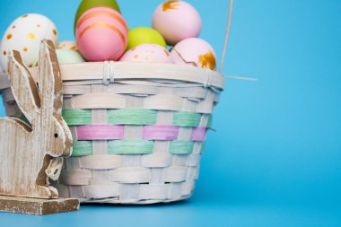 Mavi arka planda oyuncak tavşanlı bir sepet Paskalya yumurtası. Renkli boyalı yumurtalar ve oyuncak tahta tavşanla dolu bir sepet. Paskalya Kompozisyonu.