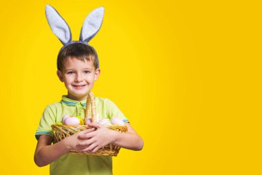 Paskalya tavşanı kulağı takan mutlu bir gülen çocuk sarı arka planda boyalı yumurtalarla dolu bir sepet tutuyor. Uzayı kopyala Paskalya Günü.