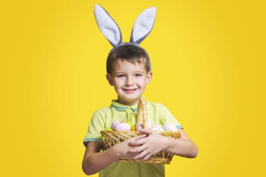 Paskalya tavşanı kulağı takan mutlu bir gülen çocuk sarı arka planda boyalı yumurtalarla dolu bir sepet tutuyor. Uzayı kopyala Paskalya Günü.