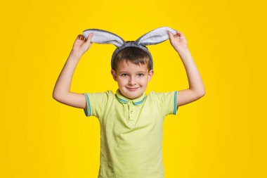 Tişörtlü küçük çocuk tavşan kulaklarına dokunuyor ve sarı arka planda dururken gülümsüyor. Paskalya günü tavşan kulağı takan şirin bir çocuk..