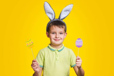 Tavşan kulaklı sevimli çocuk sarı arka planda renkli yumurtaları elinde tutuyor. Paskalya yumurtaları ve sarı arka planda tavşan kulakları olan mutlu çocuk..