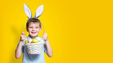 Elinde Paskalya sepeti ve kafasında sarı bir arka planda tavşan kulağı olan sevimli bir çocuk. Metin eklenecek yer ile paskalya arkaplanı.