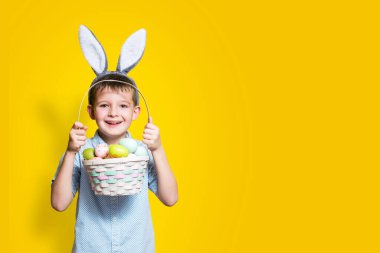 Elinde Paskalya sepeti ve kafasında sarı bir arka planda tavşan kulağı olan sevimli bir çocuk. Metin eklenecek yer ile paskalya arkaplanı.