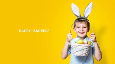 Elinde Paskalya sepeti ve kafasında sarı bir arka planda tavşan kulağı olan sevimli bir çocuk. Mutlu Paskalyalar.