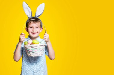 Elinde Paskalya sepeti ve kafasında sarı bir arka planda tavşan kulağı olan sevimli bir çocuk. Metin eklenecek yer ile paskalya arkaplanı.