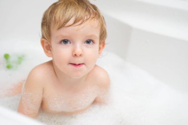 Banyoda banyo yapan komik küçük çocuk. Bir çocuk, beyaz banyoda sabun köpüğüyle suda yıkanır..