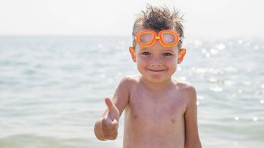 Yüzmek için gözlüklü küçük mutlu çocuk jest yapıyor. Başparmağını denizin arka planında kaldırıyor. Seyahat ve yaz tatili konsepti.