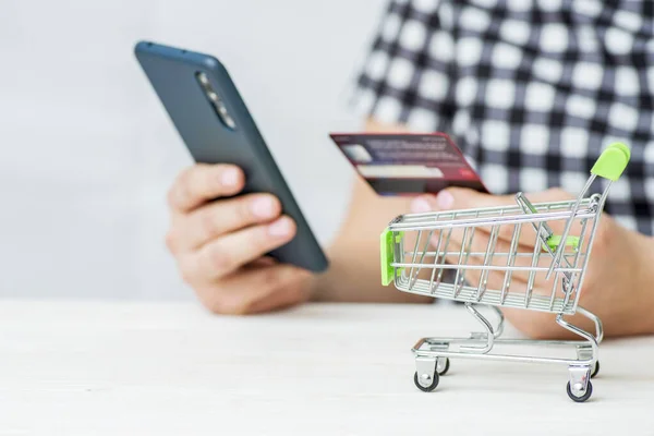 Shopping Cart and woman holding credit card and smartphone shopping online. Woman purchases goods from internet. Female makes payment on bank website.