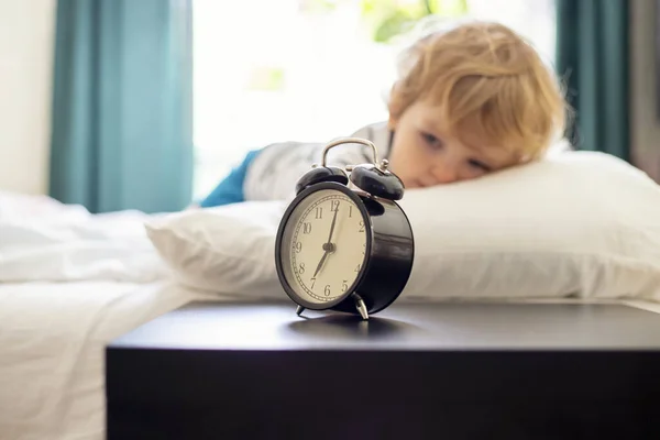 A sleepy little child lies on the bed and watches the ringing alarm clock. Time to wake up.