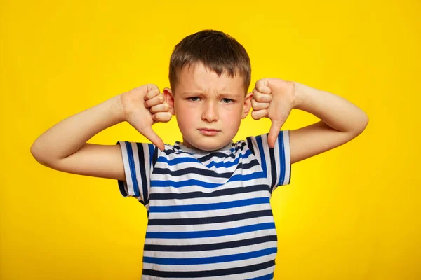 一个穿着条纹T恤的小男孩 双手插在黄色的背景上 露出大拇指垂下的姿势 不高兴的孩子把大拇指朝下用手做手势 — 图库照片
