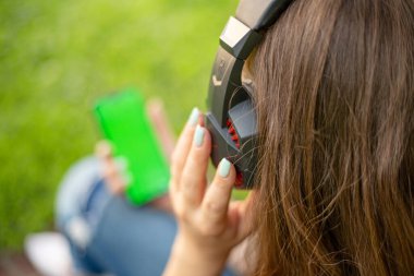 Bir kadın akıllı telefon tutuyor ve parkta çimlerin üzerinde otururken kulaklıkla müzik dinliyor..