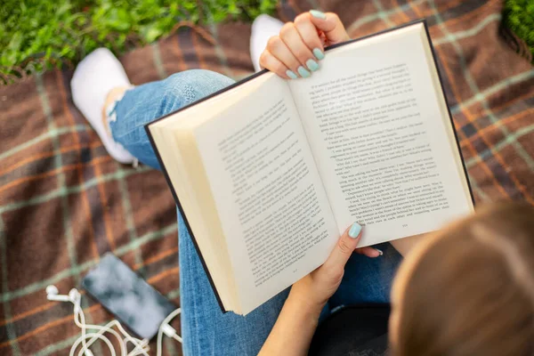 Woman in jeans with a book, a cup of coffee and a smartphone with headphones sits on a blanket in the park.