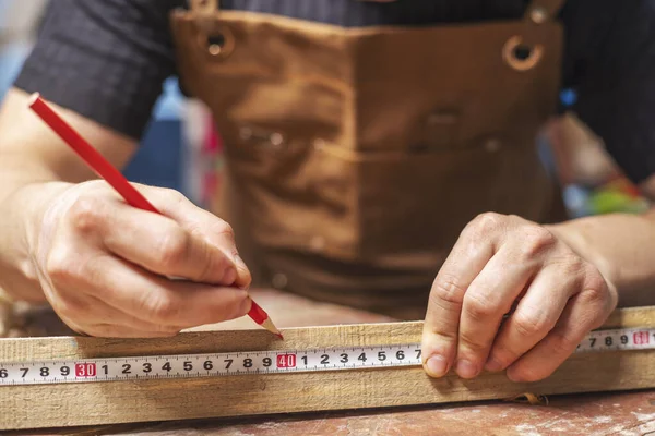 A carpenter making a mark with red pencil after measuring a wooden board with a measuring tape.