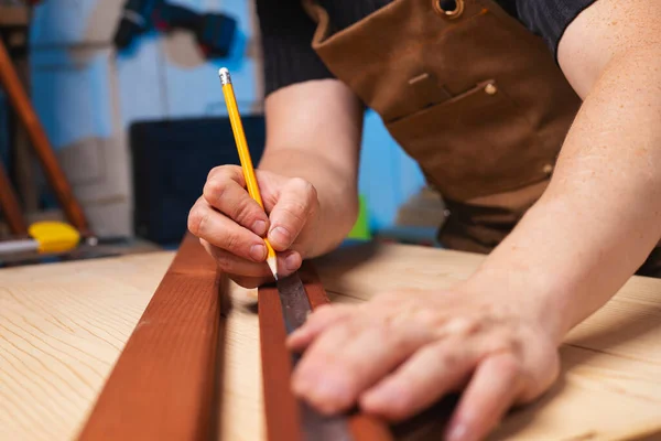 A carpenter making a mark with pencil after measuring a wooden board with a measuring tape.