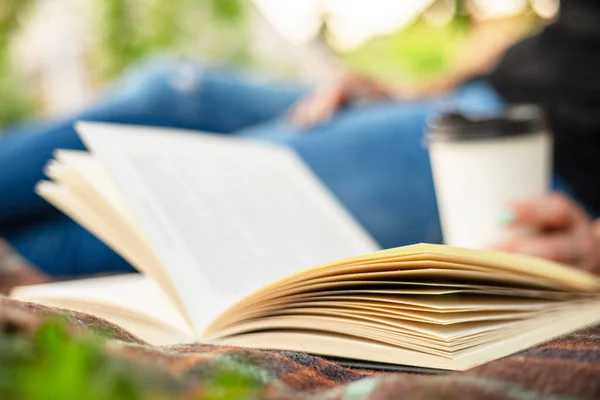 A woman in jeans with a book and a cup of coffee sits on a blanket in the park.