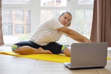 Spor giyimli güzel bir kadın evde yoga minderiyle spor egzersizi yapıyor. Bilgisayarla beden eğitimi eğitimi eğitimi. Sağlıklı yaşam tarzı kavramı.
