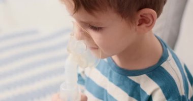 Çizgili gömlekli bir çocuk solunum terapisi için nebulizör kullanıyor. Video, çocuğun ilaçlarını soluduğu, evde sağlık ve pediatrik bakım uygulamalarını ön plana çıkardığı görüntüleri içeriyor..