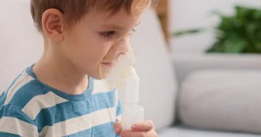 Çizgili gömlekli bir çocuk evde solunum terapisi için nebulizör kullanıyor. Olay yeri, pediatrik sağlık hizmetleri ve evde yapılan tıbbi tedavilerin önemini vurguluyor.