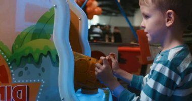 Çizgili tişörtlü genç bir çocuk kapalı bir eğlence merkezinde oyun oynuyor, dikkatle ekrana odaklanıyor. Renkli ortam neşeli atmosfere ekleniyor.