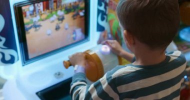 Küçük bir çocuk, bir eğlence merkezinde, ekrana dikkatle odaklanmış, ilgi çekici bir atari oyunu oynuyor. Canlı renkler ve etkileşimli kurulum heyecan verici bir oyun deneyimi yaratıyor..