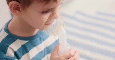 Çizgili gömlekli bir çocuk solunum tedavisi için nebulizör kullanıyor. Video, çocuğun ilaçlarını alırken, evde sağlık ve pediatrik bakımı ön plana çıkarırken görüntüleniyor..