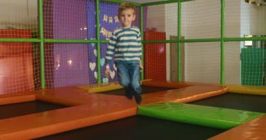 Çizgili tişörtlü bir çocuk renkli bir oyun merkezinde trambolinde enerjik bir şekilde zıplıyor. Canlı ortamda yastıklı duvarlar ve güvenlik ağları var..