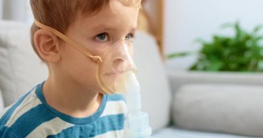 Solunum tedavisi için nebulizör kullanan küçük bir çocuk kanepede oturuyor. Sahne, pediatrik bakım ve solunum sağlığı yönetiminin vurgulandığı bir ev sağlığı ortamını yakalıyor.