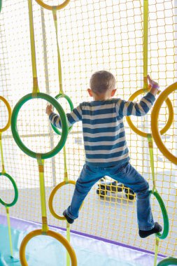 Küçük bir çocuk kapalı bir oyun bahçesinde renkli yüzüklerle ağlara tırmanıyor. Çocuk çizgili bir gömlek ve kot pantolon giyiyor, aktif oyun ve çocukluk macerası sergiliyor..
