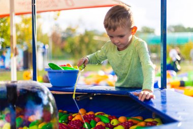Genç bir çocuk, güneşli bir günün tadını çıkararak, açık hava etkinlik istasyonunda renkli plastik meyve ve su oyuncaklarıyla oynuyor. Bu sahne çocukluk oyununun neşe ve merakını yakalar..