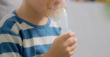 Çizgili gömlekli bir çocuk solunum terapisi için nebulizör kullanıyor. Video, çocuğun maskeyle ilacı soluyarak sağlık ve sağlığa odaklandığını gösteriyor..