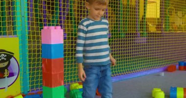 Çizgili tişörtlü küçük bir çocuk kapalı bir oyun alanında renkli yapı taşlarıyla oynuyor. Ağ örülmüş bir muhafaza ve canlı dekorasyonlar, eğlenceli bir ortam yaratıyor..
