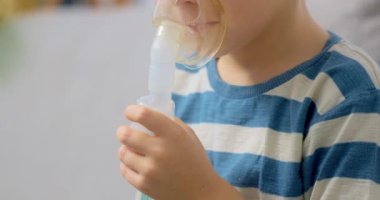 Çizgili gömlekli bir çocuk evde solunum tedavisi için nebulizör kullanıyor. Video, çocuğun maskeyle ilaç soluyarak ev sağlık hizmetlerini ön plana çıkardığını gösteriyor..