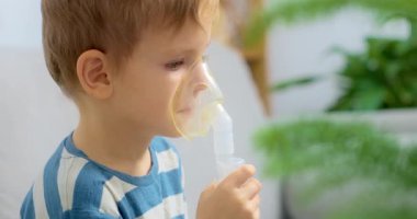 Çizgili gömlekli bir çocuk solunum tedavisi için nebulizör kullanıyor. Sahne, arka planda bitkilerin olduğu bir ev ortamında geçiyor, sağlık hizmetlerini ön plana çıkarıyor..