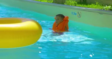 Turuncu can yeleği giyen bir çocuk su parkındaki havuzda oynar ve sarı şişme bir yüzükle etkileşime girer. Sahne neşeli ve güvenli bir günü yakalıyor..