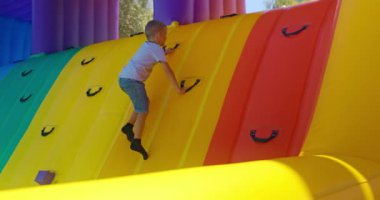 Bir çocuk açık hava çocuk parkında renkli şişme bir duvara tırmanıyor. Canlı yapı, eğlenceli ve güvenli bir tırmanış deneyimi sağlayarak destek için birden fazla kulp içeriyor..