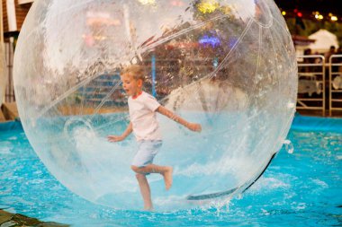 Genç bir çocuk havuzda saydam bir su topunun içinde eğleniyor. Sahne, lunaparktaki eğlence etkinliklerinin heyecanını ve eğlencesini yakalıyor..