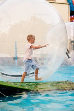 Genç bir çocuk eğlence parkında saydam şişme bir zorb topunun içinde yürüyor, eğlenceli ve benzersiz bir deneyimin tadını çıkarıyor. Sahne, neşe ve macera yakalar..