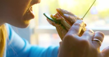 Mavi tişörtlü bir insan, bir sandviçten bir ısırık alır, güneşli bir pencerenin önünde yemeğin tadını çıkarır. Video, sandviçin detaylarını ve yeme eylemini kaydediyor..
