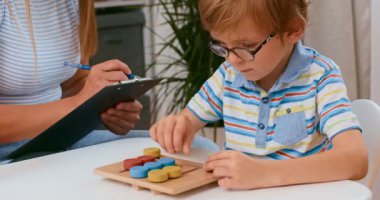 Gözlük takan genç bir çocuk renkli bir eğitim bulmacasına odaklanırken, bir öğretmen gözlemler ve notlar alır. Ortam, öğrenme ve geliştirmeyi vurgulayan bir sınıf..